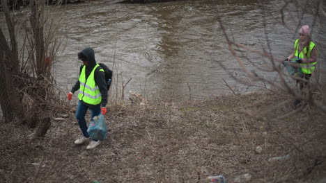 volunteer in reflective vest with gloves and backpack picking up plastic waste and trash along polluted riverbank on cold overcast day, surrounded by leafless trees during environmental cleanup effort