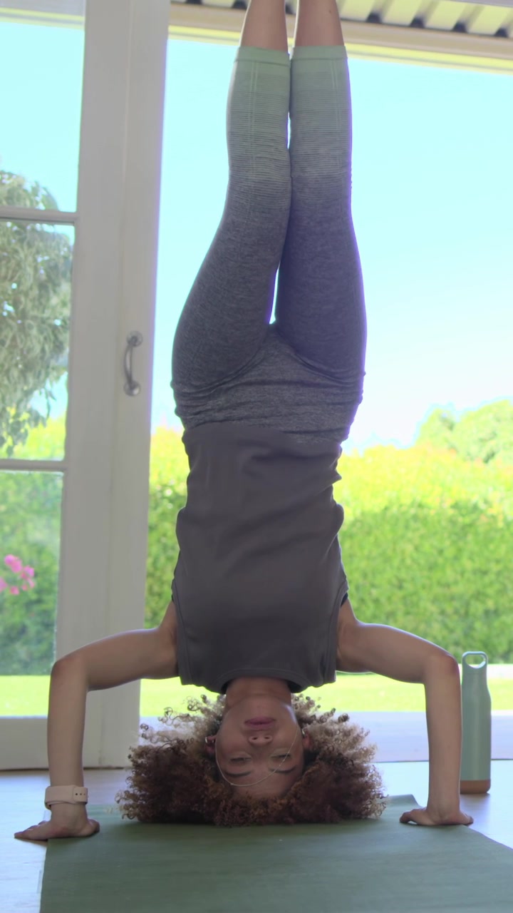 Video verticale: Donna che inizia una verticale sugli avambracci facendo yoga a casa, con attrezzatura grigia, tappetino verde e bottiglia.