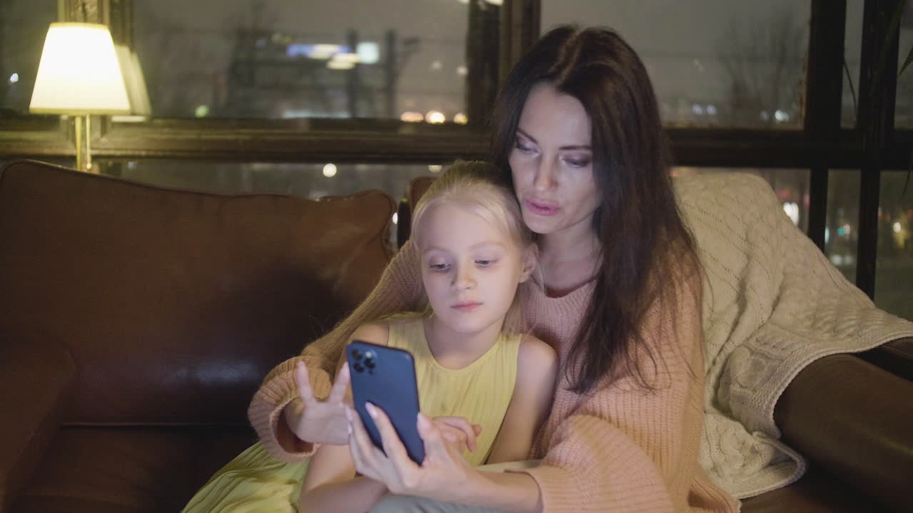 Woman Watching Something On Mobile Phone With Her Little Daughter While Sitting Together On Sofa At Home At Night