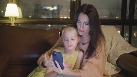 woman watching something on mobile phone with her little daughter while sitting together on sofa at home at night