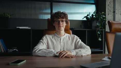 Portrait-of-a-happy-young-guy-with-curly-hair-wearing-glasses-in-a-white-shirt-who-is-sitting-at-a-table-in-the-office-and-posing