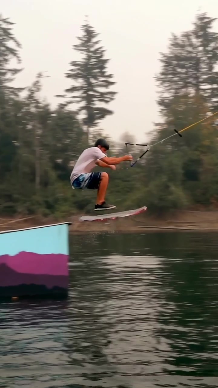Video verticale: Un wakeboarder trainato da cavo esegue carving e sale su una rampa, salta e cade (come parte dello sport), indossando un casco.