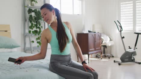 mujer asiática feliz sentada en la cama con ropa de fitness, preparándose para hacer ejercicio con un teléfono inteligente