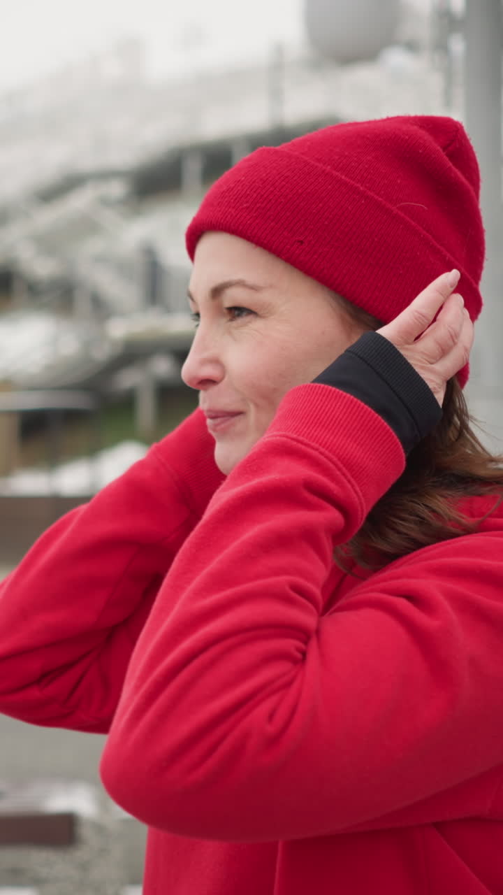 Side view of freelancer adjusting red beanie outdoors with warm smile during winter, background features snow-covered urban landscape, wooden structures, and modern benches
