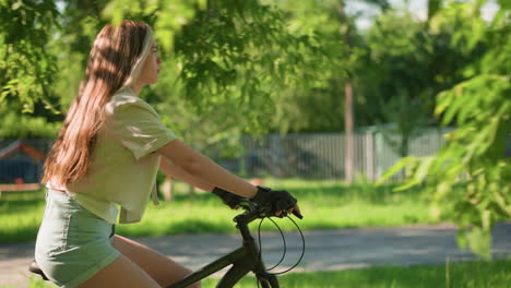 side view of young woman confidently riding bicycle along lush green park path, her long hair flowing naturally, background includes trees and iron fence