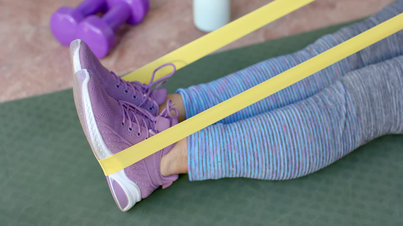 Donna anziana seduta su un tappetino verde che avvolge una fascia gialla intorno a scarpe viola, preparandosi per un esercizio per le gambe.