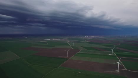 Windräder-Säumen-Eine-Weite-Grüne-Landschaft-Vor-Einem-Aufziehenden-Sturm,-Mit-Dramatischen-Dunklen-Wolken-Am-Himmel