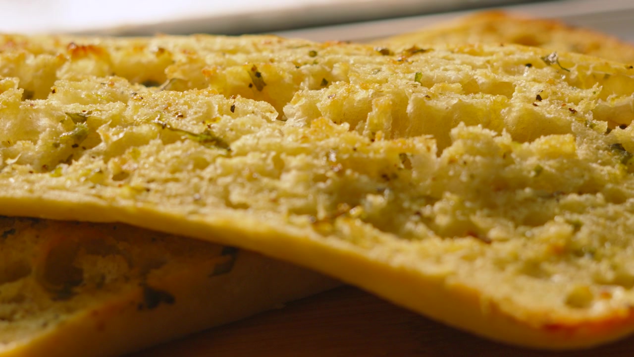 Ripresa al rallentatore di pane ciabatta artigianale all'aglio ed erbe aromatiche appena sfornato su un tagliere di legno. Perfetto per programmi di cucina, video di ricette e pubblicità di ristoranti informali