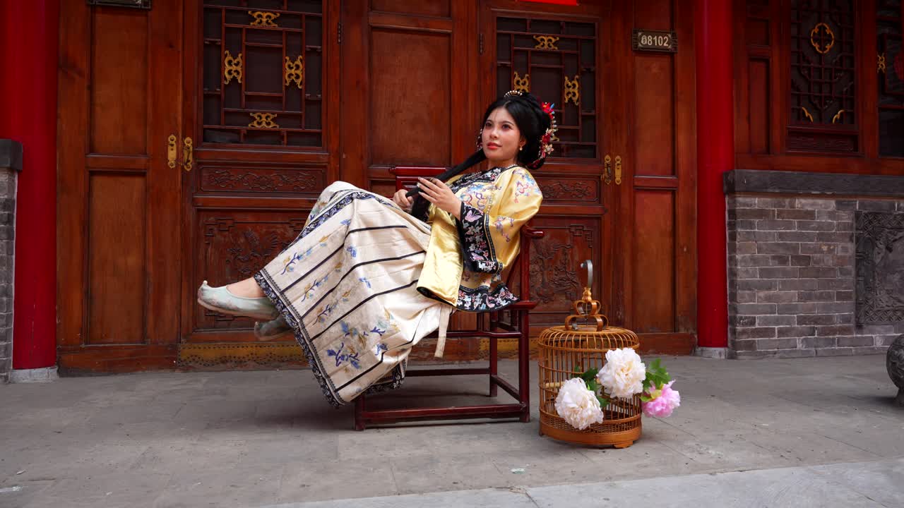 칭 왕조 여성은 머리카락으로 놀면서 미소 짓고, 전통적인 옷을 입고, 새집과 꽃이 있는 화려한 나무 문 에 앉아 의자에 우아하게 앉아 있습니다.