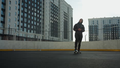 man in urban sports arena balances on one leg on soccer ball, hands clasped, turning head, high-rise buildings in background under clear sky