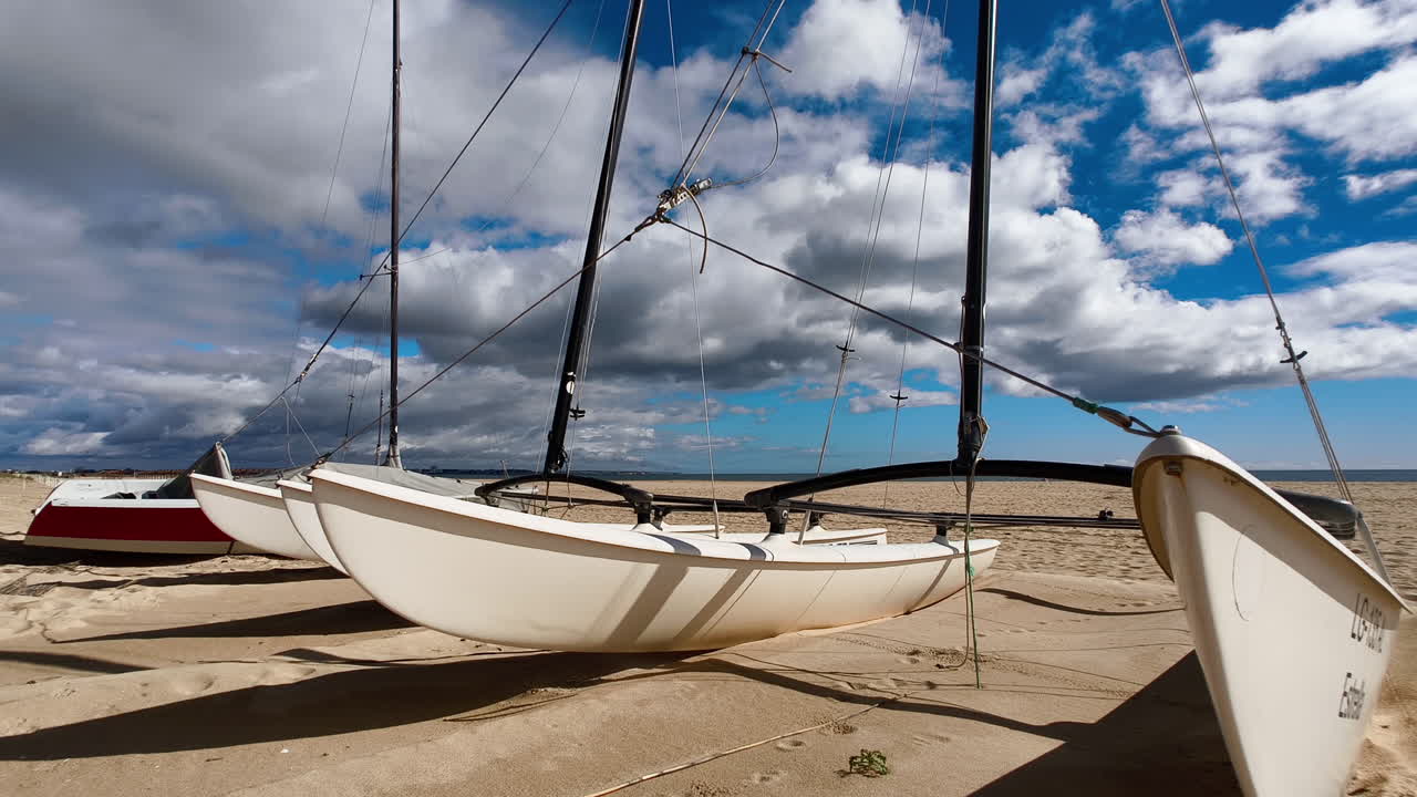 A group of Catamarans on the beach, with its sails furled. The sky is a bright blue with fluffy white clouds.In Algarve, Portugal.