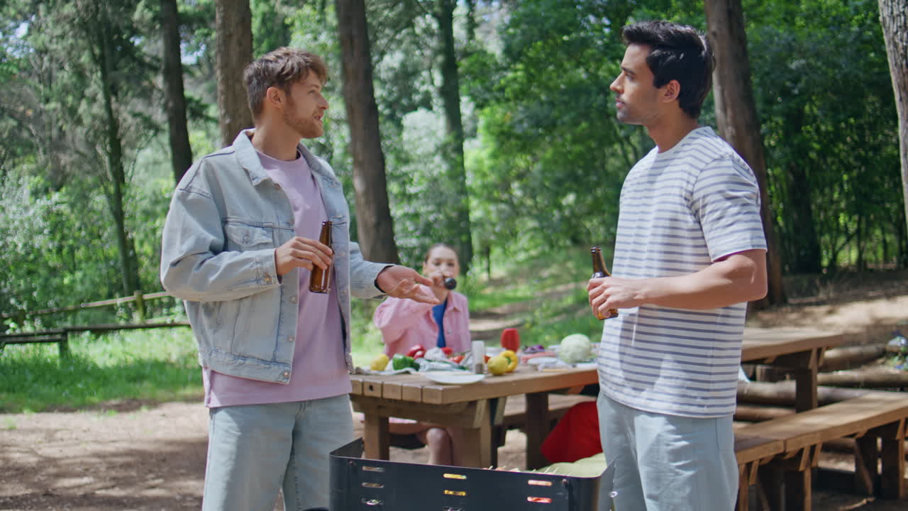 Friends enjoy grilling meal drinking beer in green forest. Smiling men talking