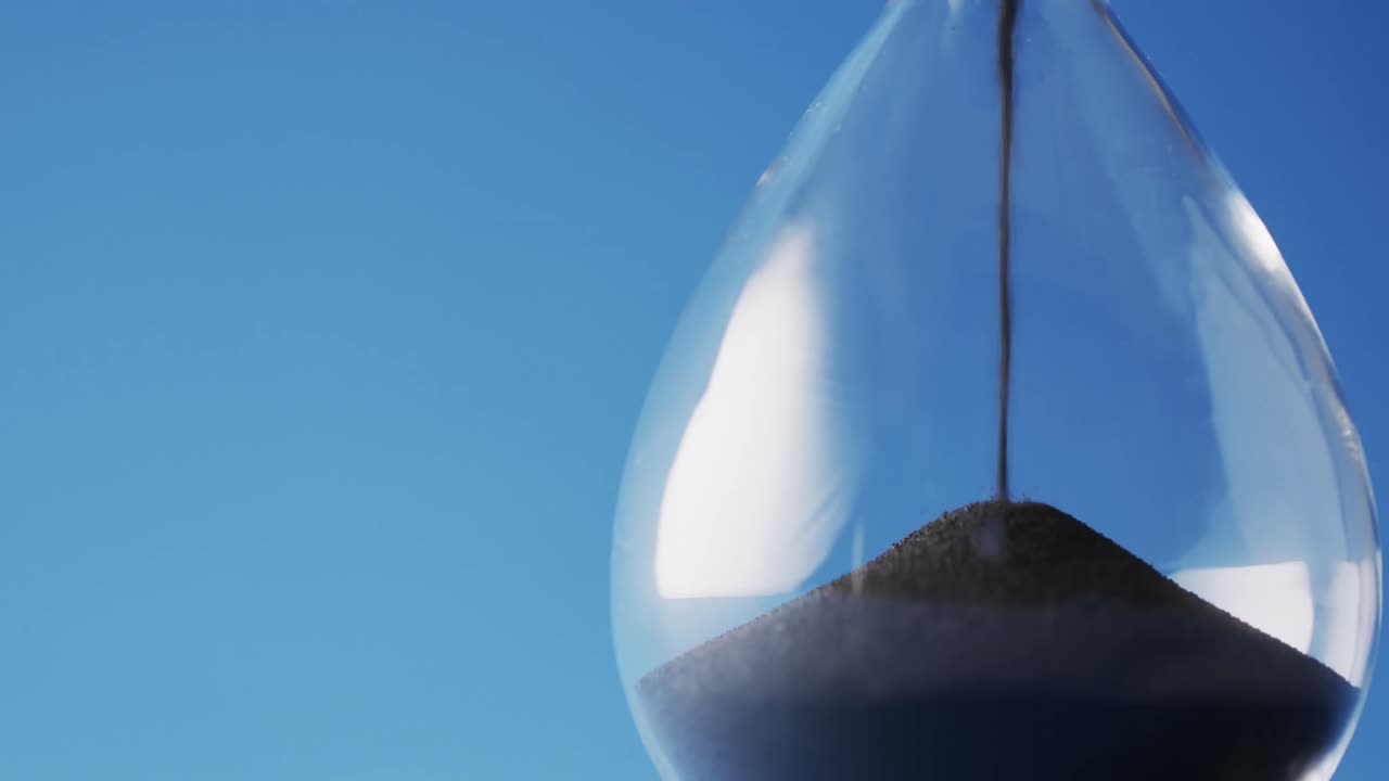 Video of close up of hourglass with black sand pouring with copy space on blue background