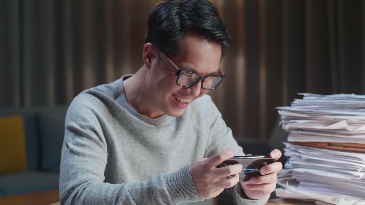 집에서 문서 작업을 한 후 스마트폰에서 게임을 잃은 아시아 남성이 화를 내고 있습니다.