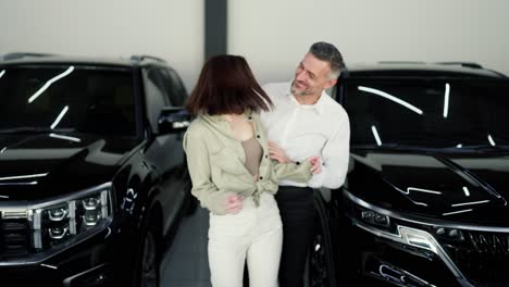 happy middle-aged man opens his girlfriend eyes with his brunette hands while informing her about buying a modern car at a car dealership. happy brunette girl receives a surprise while buying a car at a car dealership