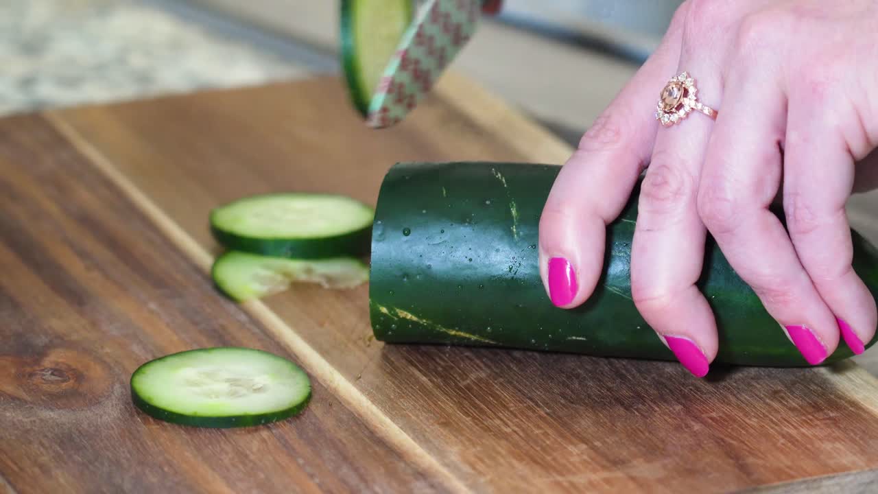 mujer cortando pepino en rodajas finas de cerca en una tabla de cortar de madera con un cuchillo pequeño decorado con flores, manos con esmalte de uñas rosa