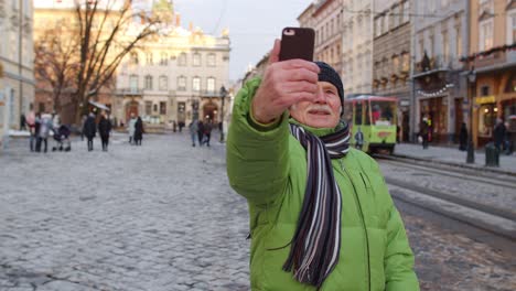 Elderly-tourist-grandfather-traveling,-taking-selfie,-making-online-video-call-on-winter-city-center