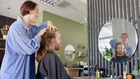 happy caucasian female hairdresser advising male client with long hair at salon, in slow motion