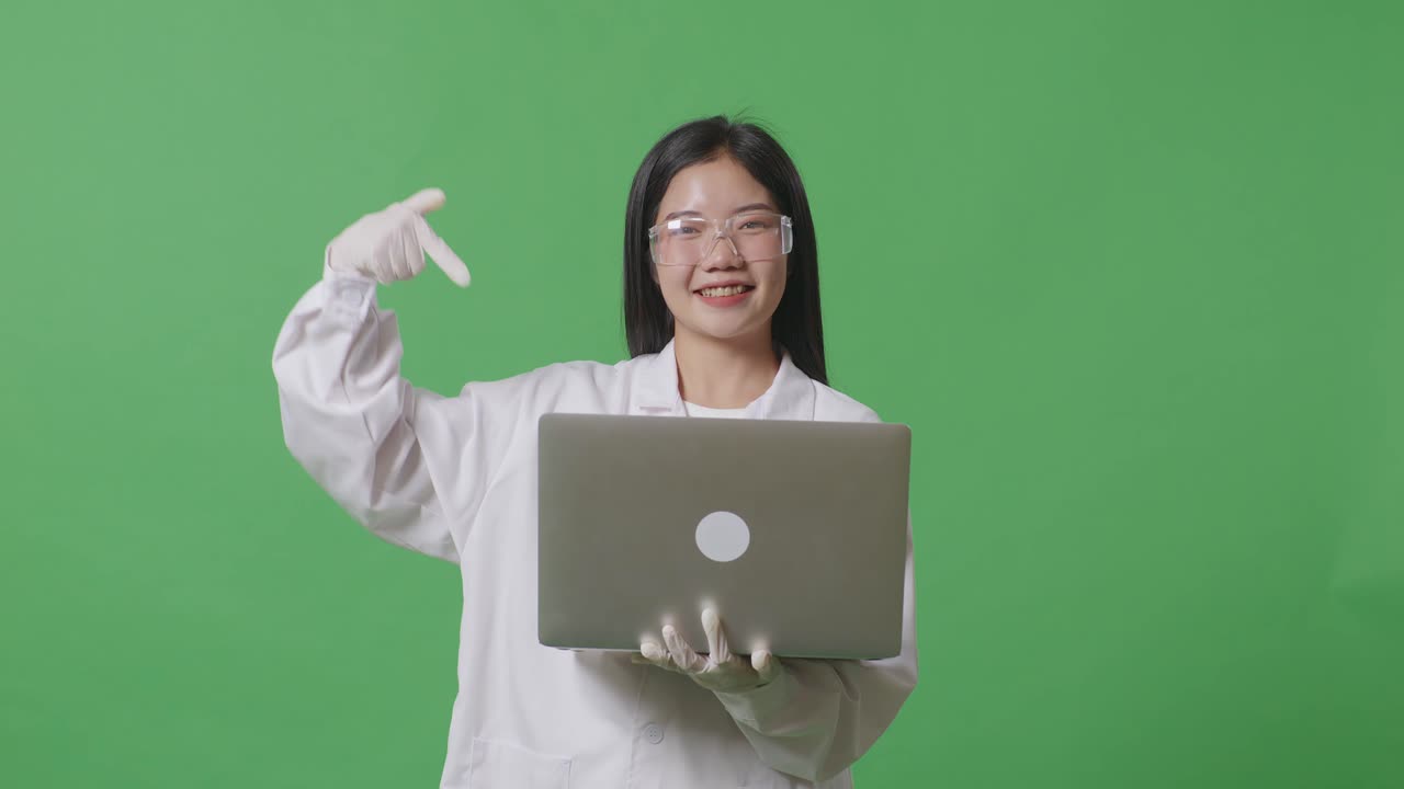 아시아 여성 과학자가 미소 짓고 노트북을 가리키며 실험실의 녹색 화면 배경에 서 있습니다.