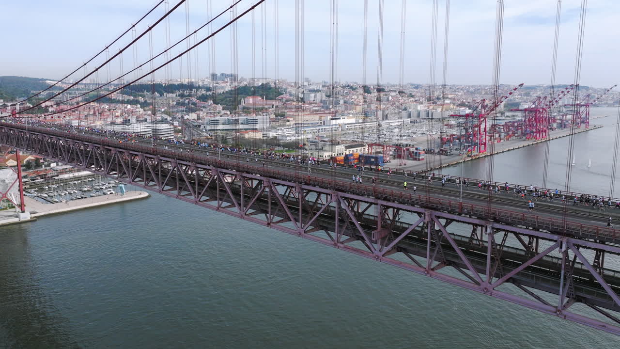 Half marathon and 10K long distance running event in Lisbon, Portugal, Europe. Runners crossing the iconic famous red 25th April suspension bridge. Aerial drone shot facing the Alcantara docks