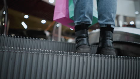 vista inferior del cuerpo de una mujer con botas negras y vaqueros llevando bolsas de compras mientras sale de una escalera mecánica en un vibrante centro comercial, mostrando movimiento, moda y experiencia de compras urbanas