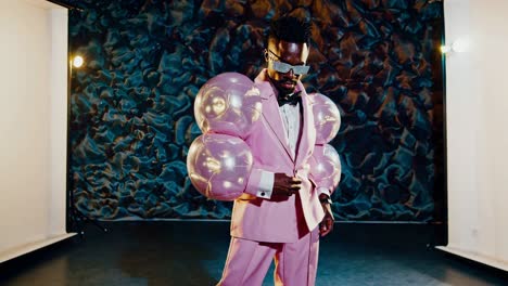 confident male model wearing vibrant pink suit featuring balloon adorned sleeves, sporting designer sunglasses while striking sophisticated pose against dramatic dark studio background