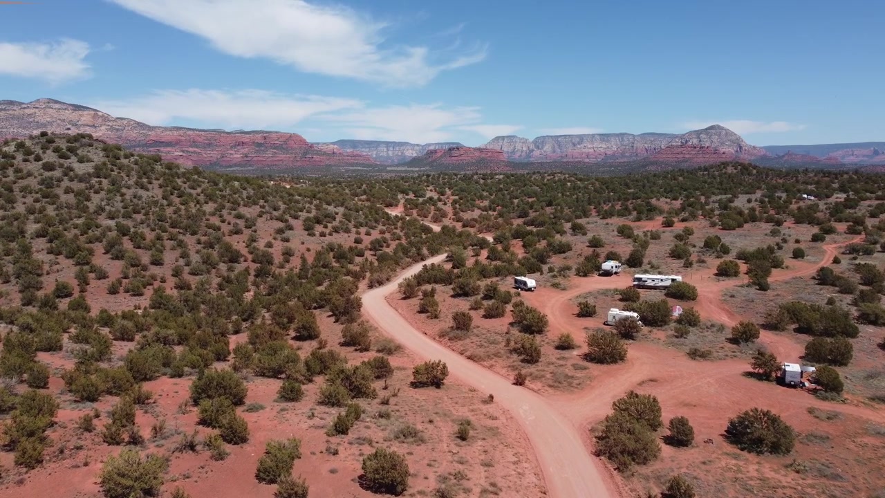 Arizona, Sedona'nın çöl manzarasında, toprak bir yol ve serbest kamp yapan karavanları gösteren drone havadan görünümü. Arka planda berrak mavi gökyüzünün altında görkemli kızıl kaya platoları ve dağlar yükseliyor.