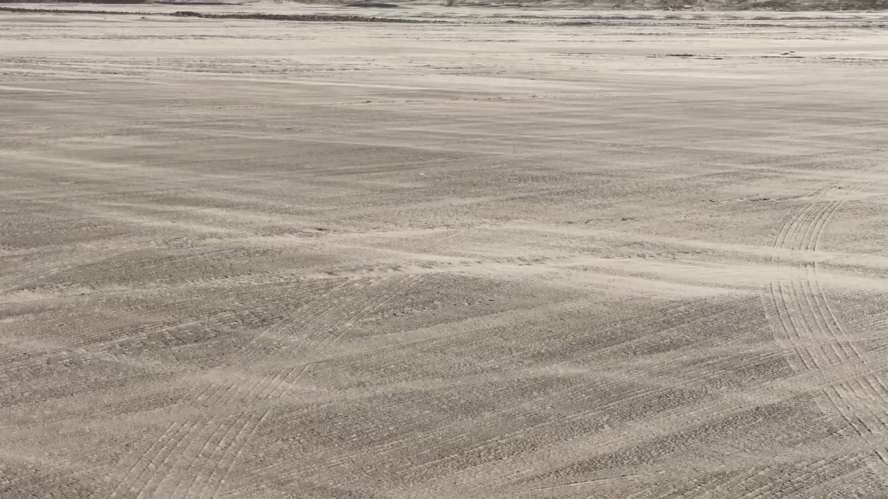 Statisk udsigt over vind, der skaber flydende sandmønstre på en flad sandstrand.
