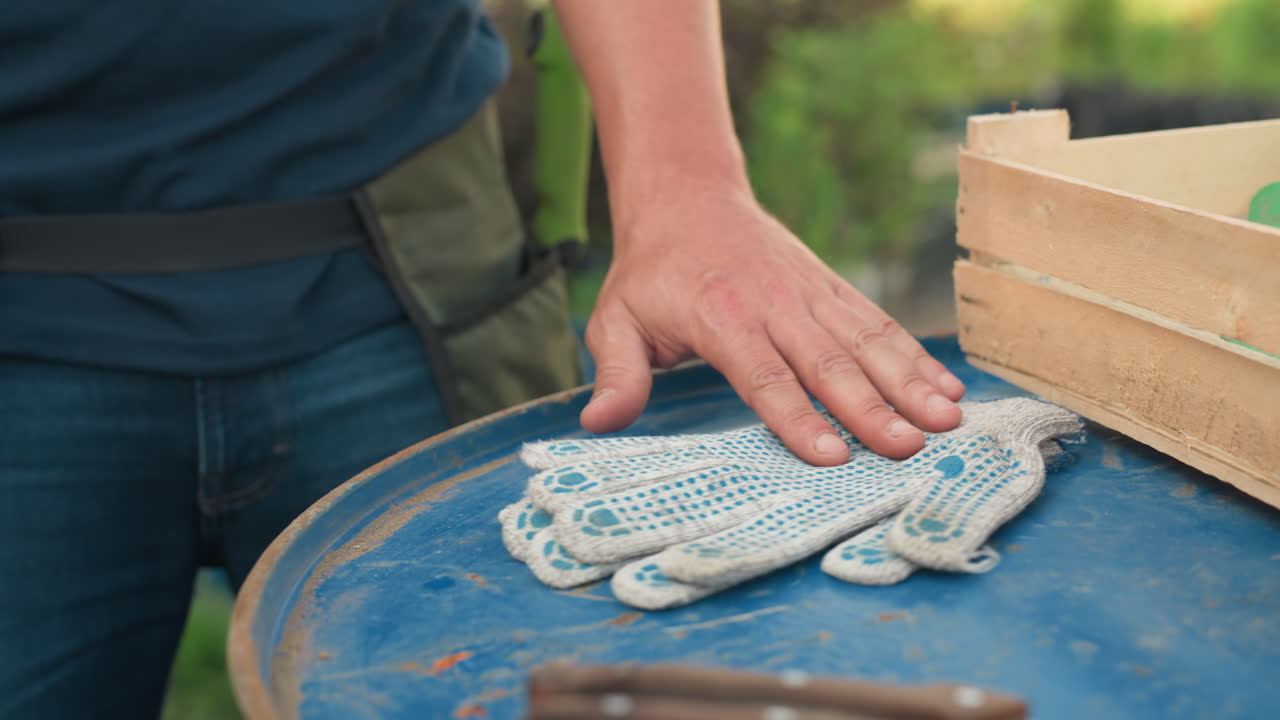 Male gardener in work apron drops work gloves onto blue metal drum beside wooden crate, hands offload gear before task, close view outdoors in plant nursery