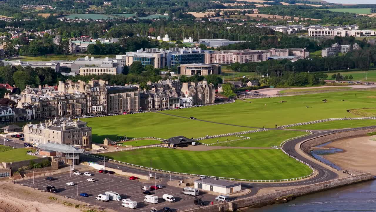 Drone captures sweeping daytime panorama of golf course, historic buildings, coastline, and parked cars
