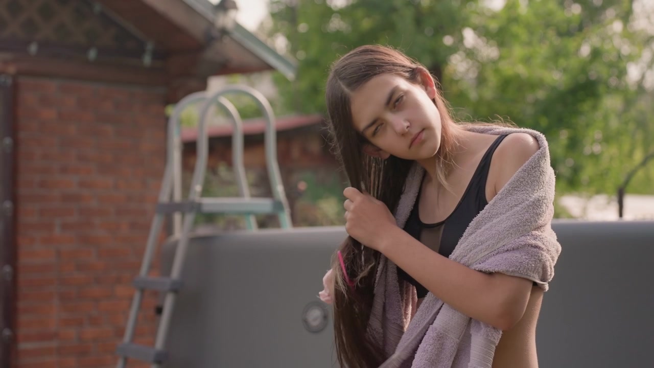 Una giovane donna si asciuga e si pettina i capelli all'aperto vicino a una piscina, indossando un reggiseno sportivo nero con un asciugamano appoggiato su di lei. L'ambiente include vegetazione e parte di un giardino visibile in una luce soffusa.