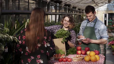 Cheerful-saleswoman-with-her-husband-is-putting-fresh-parsley-in-paper-bag-to-customer-in-greenhouse-market-talking-smiling