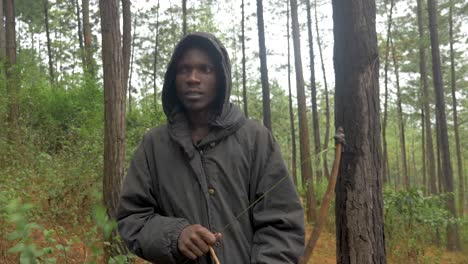 a slow motion shot tracking from in front an african hunter wearing a hooded coat as he walks through a forest hunting with a bow and arrow