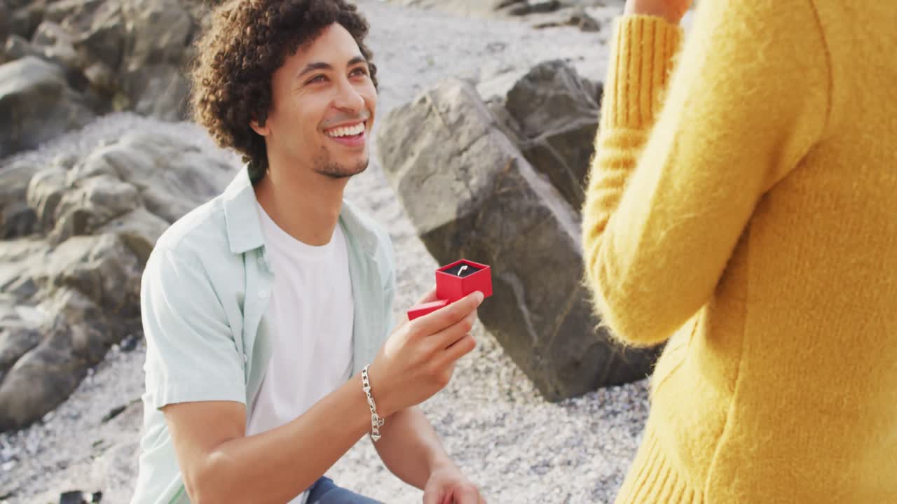 아프리카계 미국인 남자가 바다 근처의 바위 위에 반지를 들고 여자친구를 제안하고 있습니다.