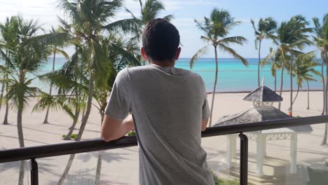 Un-Hombre-Adulto-Joven-Tiene-Vistas-A-La-Playa-Caribeña-Desde-El-Balcón-De-La-Habitación.