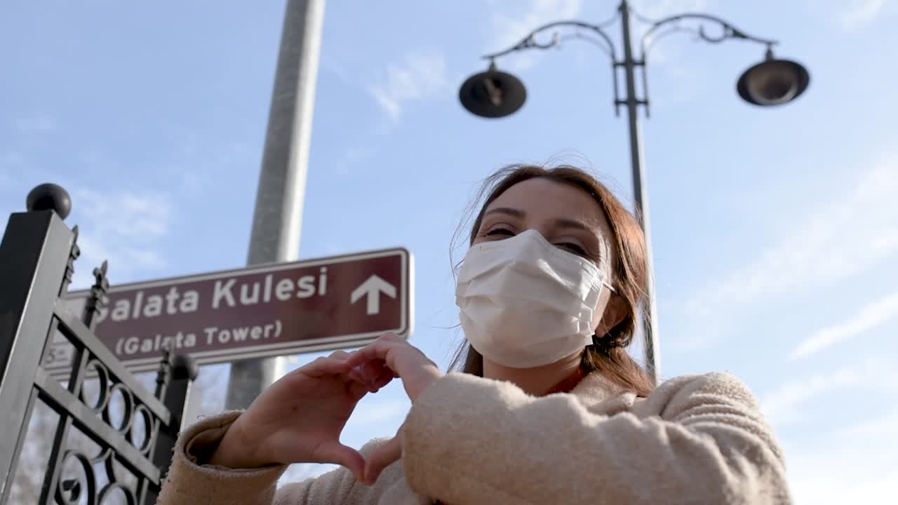 보호용 의료용 마스크와 세련된 옷을 입은 아름다운 소녀는 갈라타 타워의 새로운 정상적인 생활 방식, 여행 개념의 징후를 보여줍니다