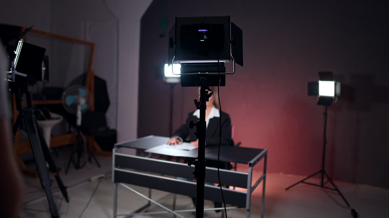 Female reporter in black jacket sits in the photo studio. Silhouette of a lady standing backstage.