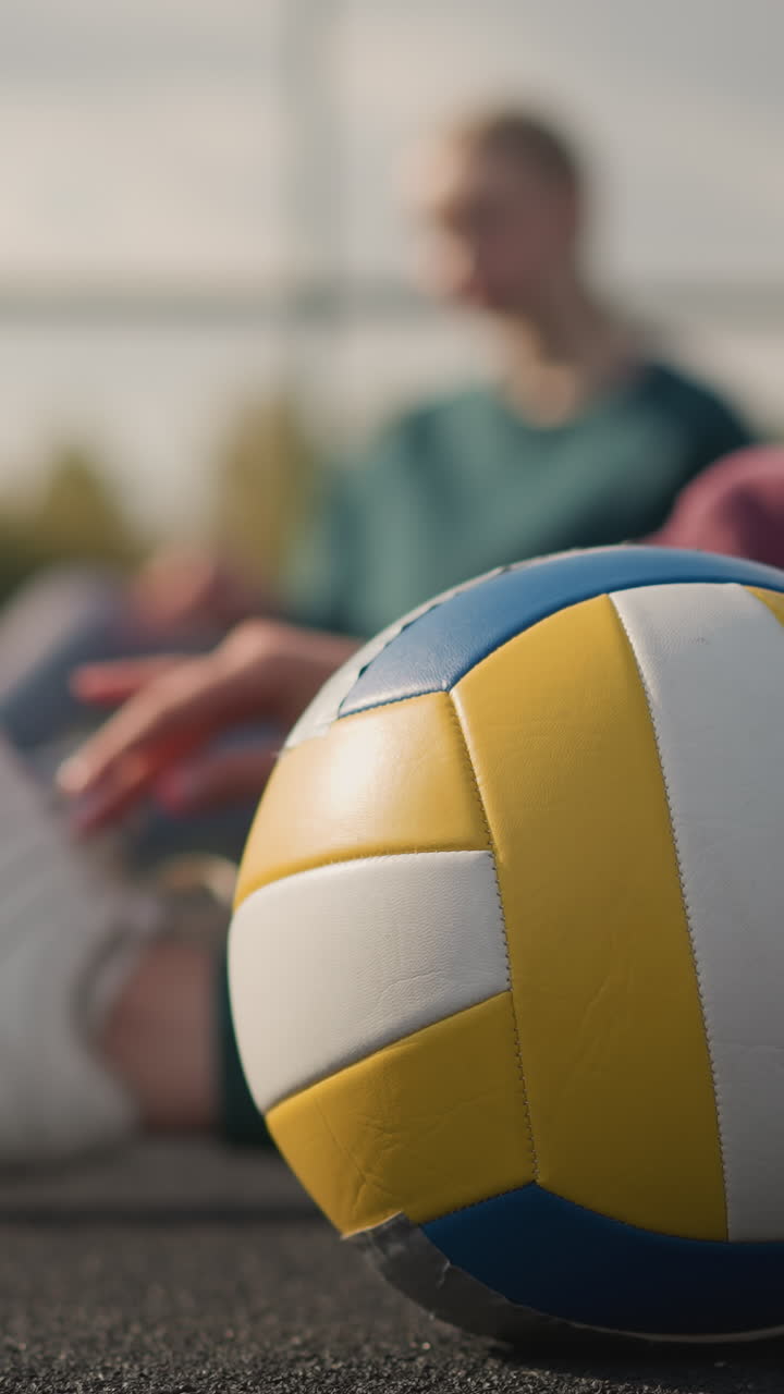 Close-up of a volleyball under someone's foot on an outdoor court with a blurred view of a woman stretching in the background, the scene captures a workout session in a sports training environment