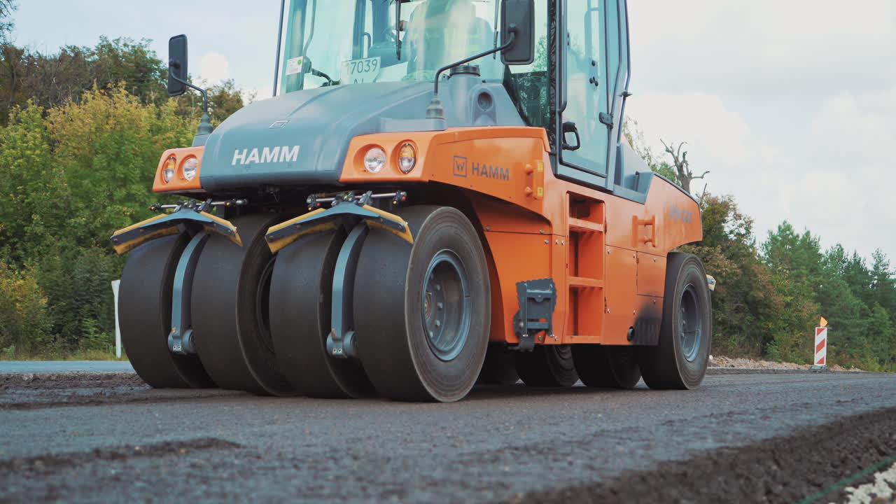 Close-up of compactor machine at work on the road. Heavy paver machine is pressing hot asphalt outdoors on green trees background.