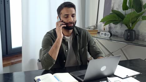 hombre positivo indio o árabe, vestido con ropa elegante, sentado en su oficina moderna, hablando por teléfono móvil con un amigo o cliente mientras trabaja en una computadora portátil, teniendo negociaciones amistosas