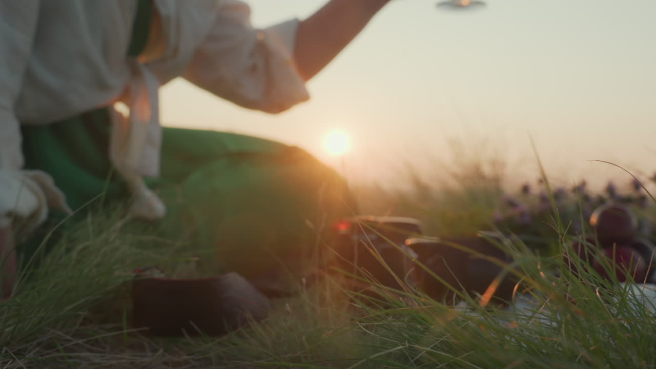Close up grass blades in foreground with blurred woman seated on hilltop holding wine glass near picnic flowers and camera, sipping red wine and gazing at distant sunset over river in golden hour