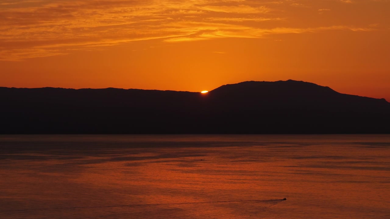Luftfoto fra drone af solnedgangen bag et bjergrigt forbjerg over havet, med den orange himmel spejlet i det rolige vand, Kreta, Grækenland