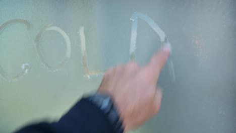 man's finger writes the word cold on window frost on brisk morning