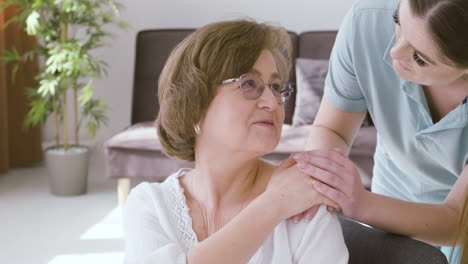 senior woman sitting on a sofa talking to a female doctor who puts her hand on her shoulder