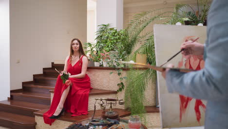 mujer con vestido de hendidura posa para el pintor en la galería. elegante dama rica en vestido rojo se sienta en el paso en el estudio del artista en el dibujo en vivo. proceso de creación de retrato
