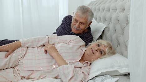 Senior-Caucasian-grandparents-couple-lying-in-bed-at-home-in-morning.-Man-gently-whispering-to-woman