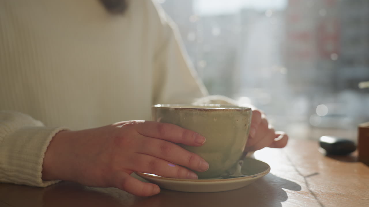Close up partial view of person holding ceramic tea cup with both hands on wooden table, soft natural light, background features moving car and blurred winter cityscape outside window
