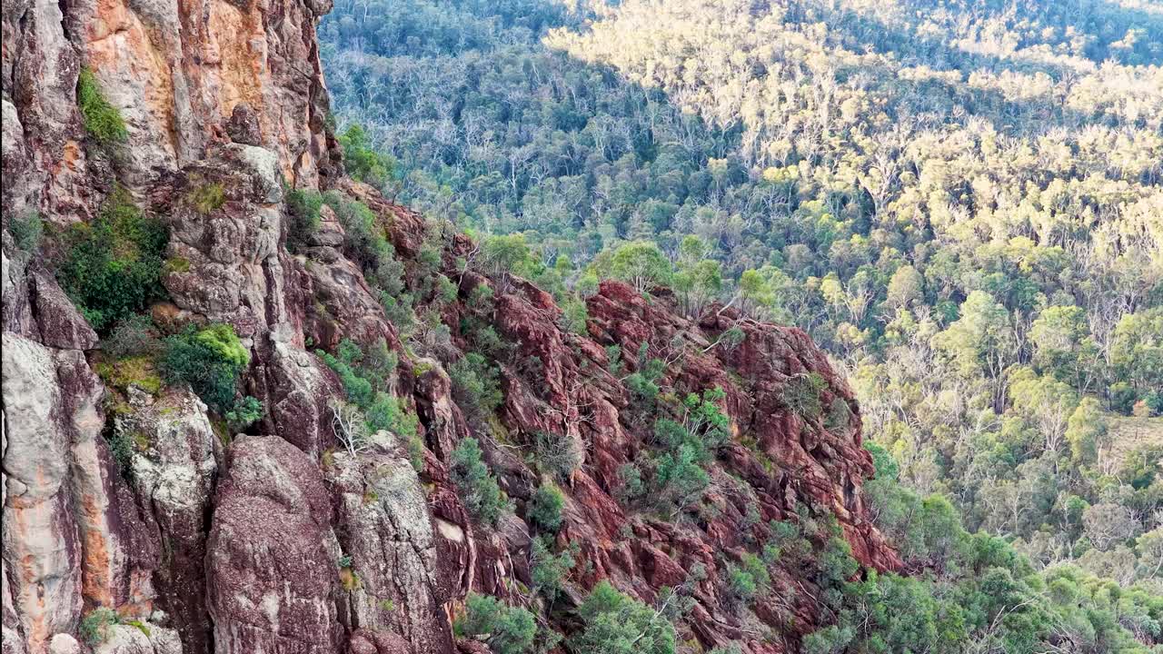 Drone pans across rugged volcanic outcrop and forested landscape in soft sunset lighting
