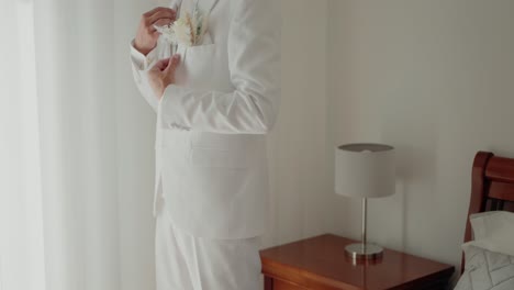 groom in a white suit adjusts his floral boutonniere while standing indoors near a bedside table, preparing for the wedding