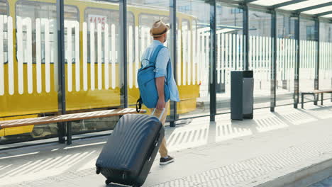 Rear-view-of-Caucasian-traveller-wearing-hat-with-backpack-and-carrying-suitcase-in-the-bus-stop